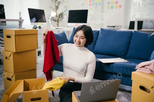 Preview: Asian female entrepreneur showing clothes in a live stream using laptop at home office