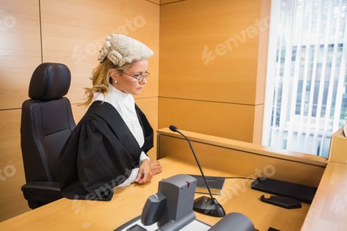 Preview: Female judge in wig, robe and glasses reviewing files in courtroom with microphone, copy space