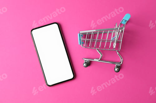 Preview: Phone with empty screen and shopping cart on pink background, top view