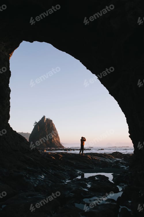 Preview: Man standing beneath sea cave at dusk, using binoculars at Hole-in-the-Rock, Rialto Beach, Olympic