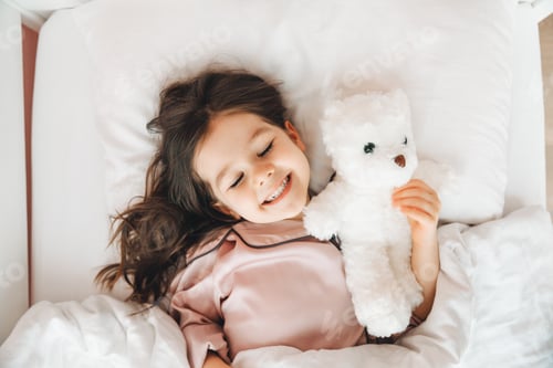 Preview: a little dark-haired girl is sleeping in bed, hugging a teddy bear under a white blanket.the child