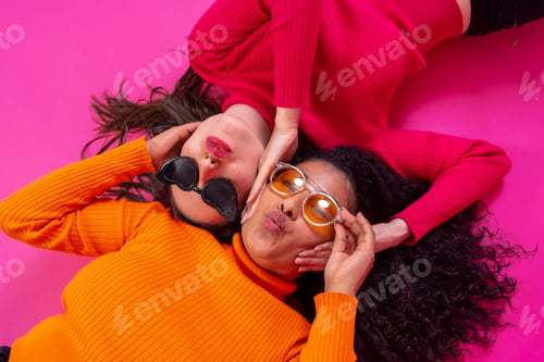 Preview: Two multi-ethnic women in sunglasses lying down smiling on a pink background, lifestyle