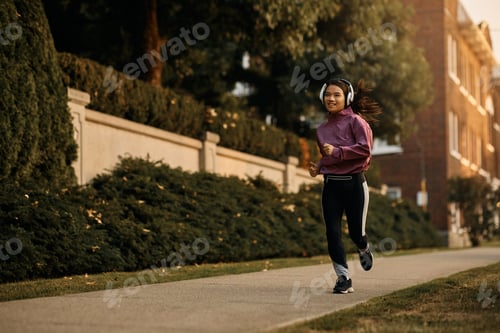 Preview: Happy Asian athletic woman listening music on headphones while running on the street.