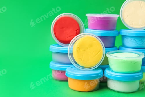 Preview: Different colorful modeling clay in plastic jars on green background, space for text