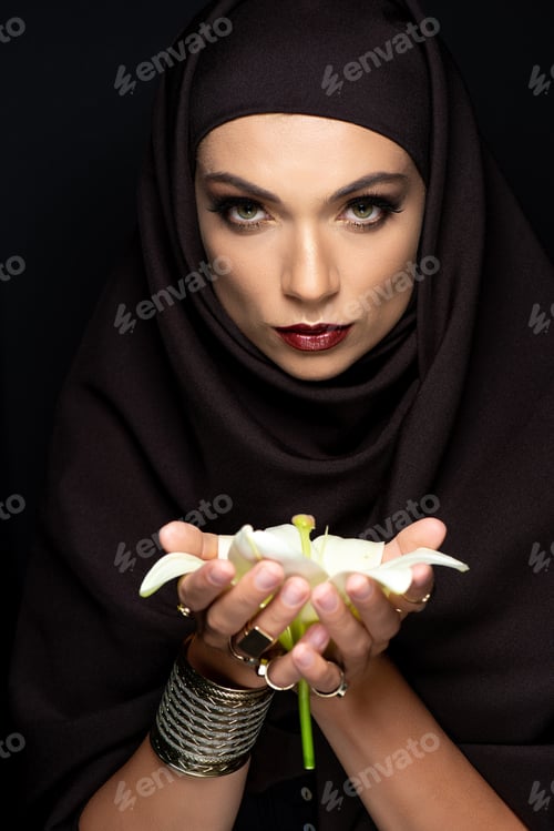 Preview: beautiful Muslim woman in hijab in golden rings and bracelet holding lily isolated on black