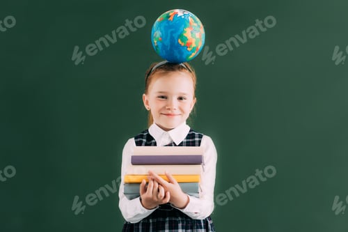 Preview: little redhead schoolgirl with globe on head holding pile of books and smiling at camera