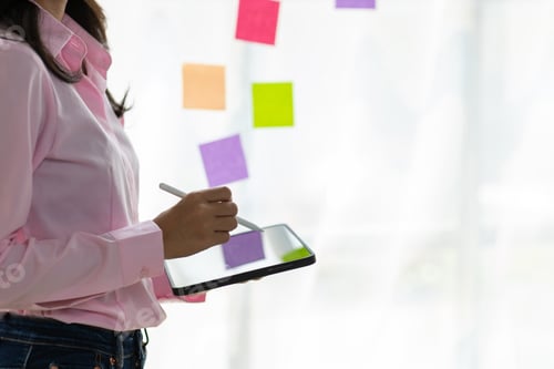 Preview: Businesswoman holding a tablet is planning her work with paper stickers on a glass plate.