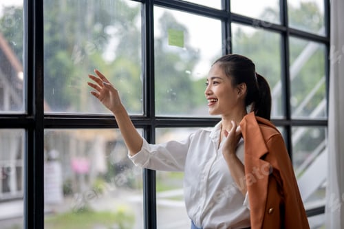 Preview: Happy businesswoman looking out of office window touching glass with hand