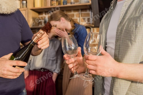 Preview: cropped shot of men holding bottle of wine and wine glasses while women gossiping behind at home
