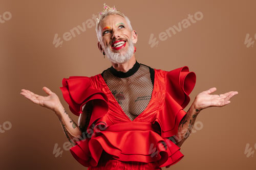 Preview: Happy gay man with crown on head wearing beautiful red dress and gesturing against brown background