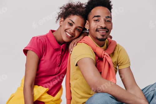 Preview: Dark-skinned guy and girl sitting on white background. Joyful woman in red shirt and brunette man i