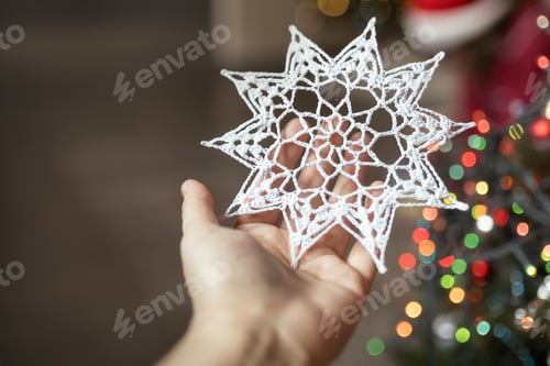 Preview: knitted white handmade snowflake with ornaments in hand on background of Christmas tree at home
