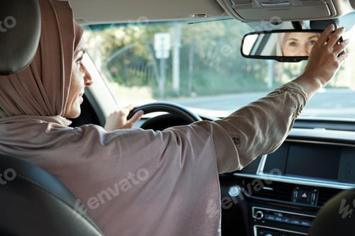 Preview: Young woman in hijab looking in mirror while driving car