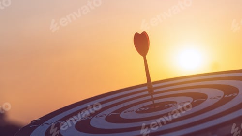 Preview: Close up red dart arrow hitting target center dartboard on sunset background.