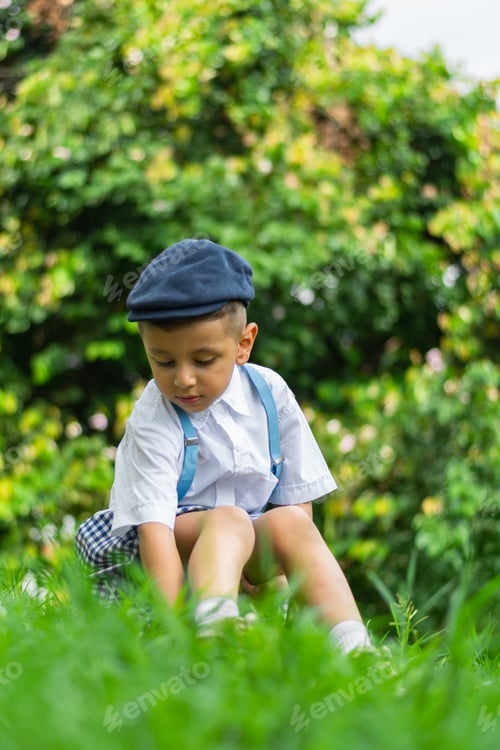 Preview: Latino boy sitting on the ground in the park, looking at his shoes, surrounded by green nature.