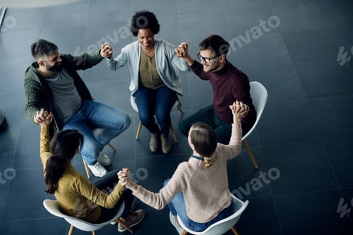 Preview: Happy people holding hands while sitting in a circle during group therapy meeting.