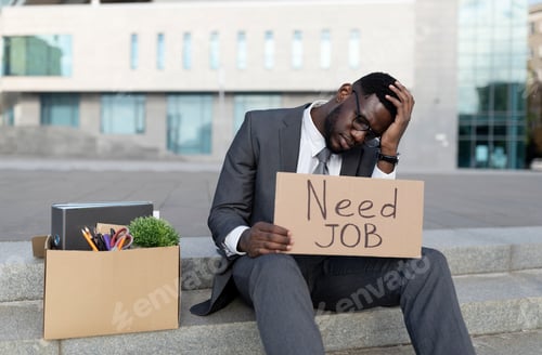Preview: Unhappy black businessman sitting outdoors with box of office supplies and poster with need job text