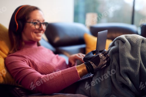 Preview: Woman With Prosthetic Arm Wearing Wireless Headphones And Working On Laptop On Sofa At Home