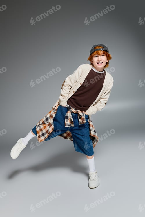Preview: Trendy redhead boy showcases stylish outfit in modern studio setting with playful pose