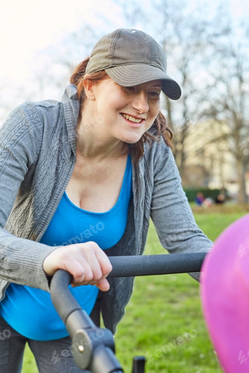 Preview: Pregnant woman wearing baseball cap looking at baby in buggy