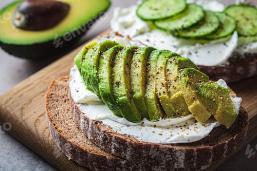 Preview: Toasts with cream cheese, avocado, cucumber and rye bread on wooden board, dark background.
