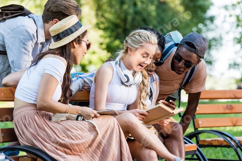 Preview: multicultural group of students reading book while sitting on bench together in park