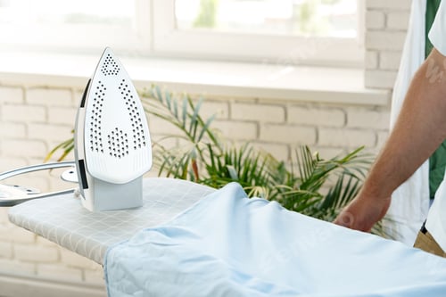 Vista previa: Ironing a light blue shirt on an ironing board with a sunny window background