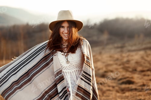 Preview: beautiful boho woman hipster having fun, wearing hat and poncho at sunset in mountains