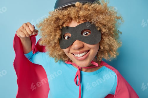 Preview: Brave cheerful young woman with curly hair clenches fist wears black eyemask costume with cloak pret
