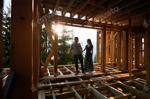 Preview: Man and woman inspecting their future wooden frame house nestled in the mountains near forest.