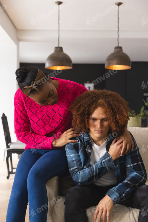 Preview: Thoughtful, sad diverse couple sitting in living room at home, embracing and comforting