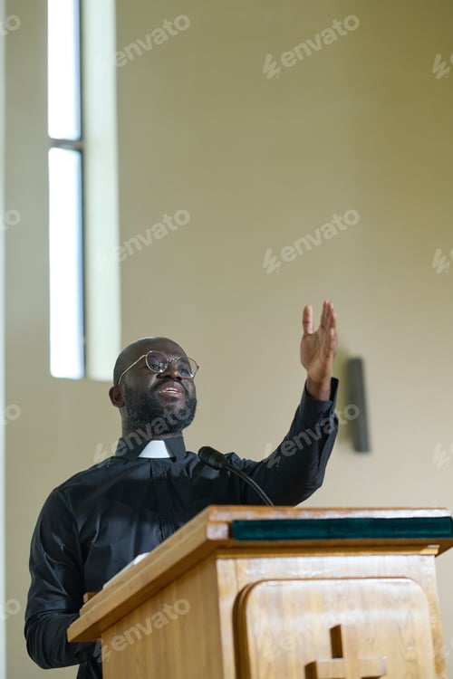 Preview: Young confident priest in black shirt with clerical collar standing by pulpit