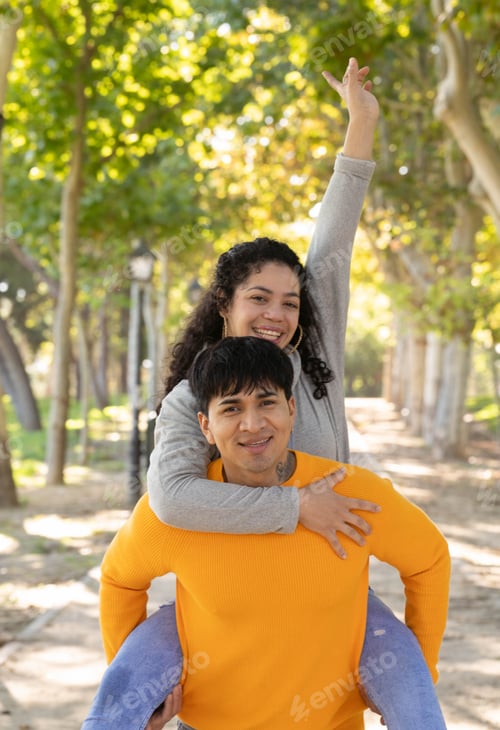 Preview: Portrait of hispanic couple in piggyback outdoors in a park raising a hand as victory or success