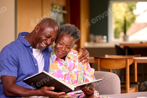 Preview: Smiling Senior Couple Sitting On Sofa At Home Looking Through Photo Album Together