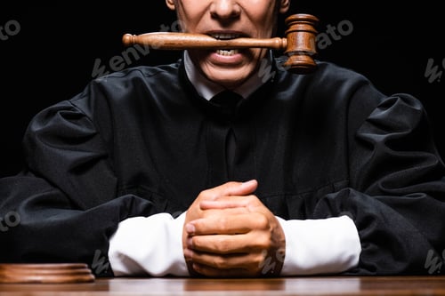Preview: cropped view of judge in judicial robe sitting at table and holding gavel in mouth isolated on black