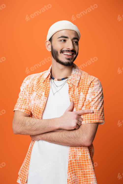 Preview: fashionable bearded man smiling and pointing with finger while looking away isolated on orange