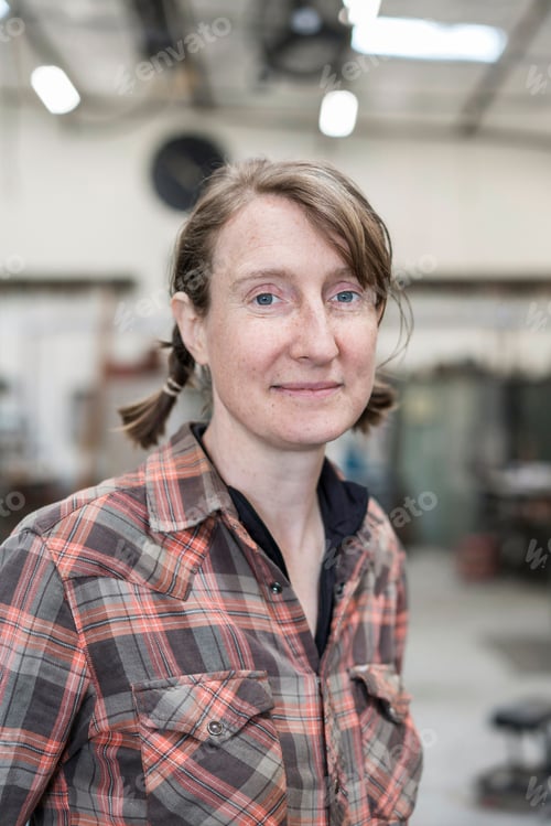 Preview: Blond woman wearing checkered shirt standing in metal workshop, smiling at camera.