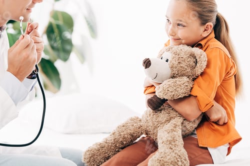 Preview: partial view of pediatrist in white coat and kid with teddy bear in clinic