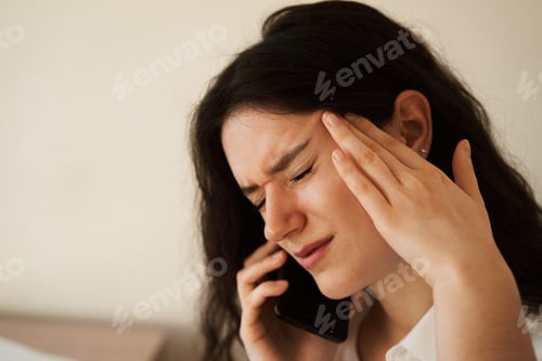 Preview: Exhausted girl with headache talks on phone, close-up portrait at home.