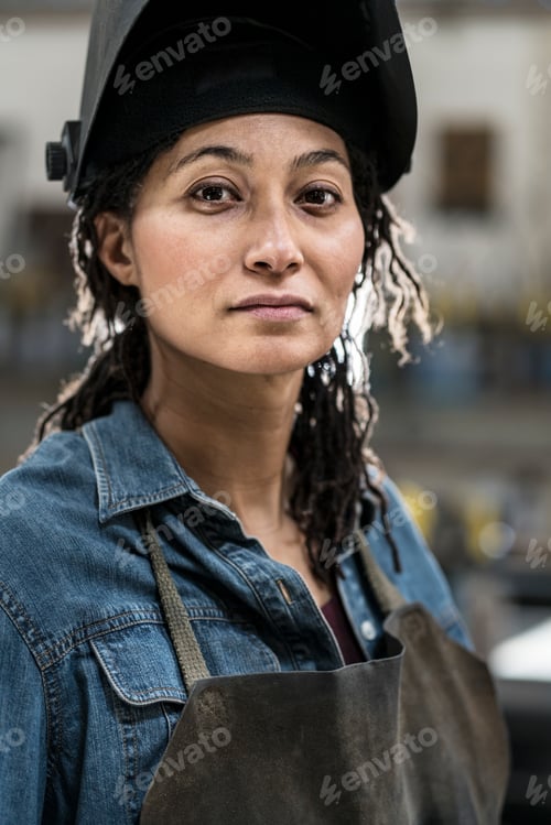 Preview: Portrait of woman wearing apron and welding mask standing in metal workshop, smiling at camera.