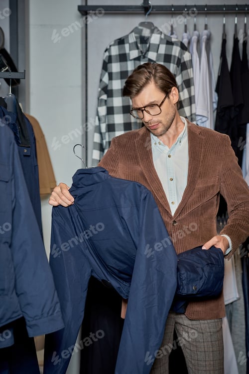 Preview: handsome man in eyeglasses holding jacket while shopping in boutique
