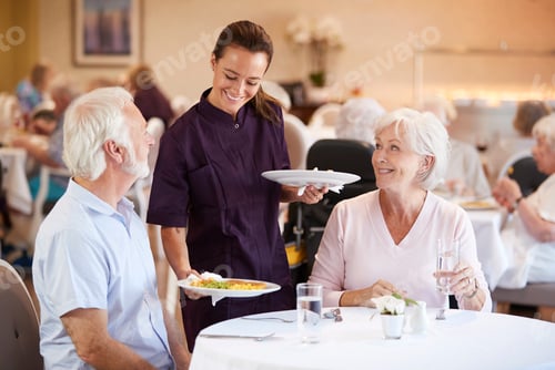 Preview: Senior Couple Being Served With Meal By Carer In Dining Room Of Retirement Home