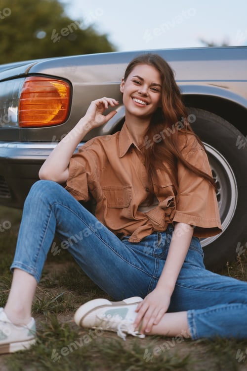 Preview: Young woman smiling in casual outfit sitting near vintage car surrounded by nature, conveying