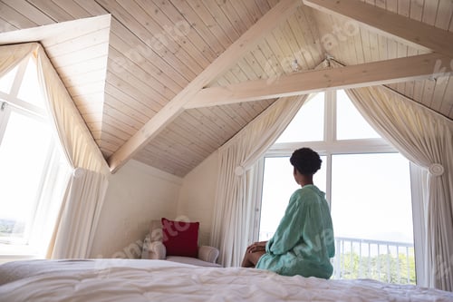 Preview: Woman looking out of the window while sitting on her bed