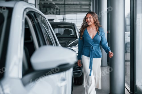 Preview: Positive woman in blue shirt walks near brand new car. In auto salon