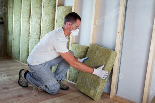 Preview: Worker in protective goggles and respirator insulating rock wool insulation in wooden frame