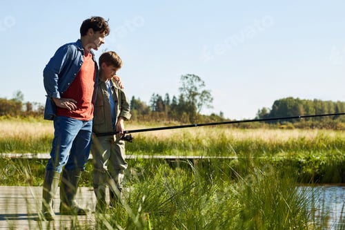 Preview: Caucasian Young Adult Man Guiding Caucasian Boy Fishing on Lakeside Dock