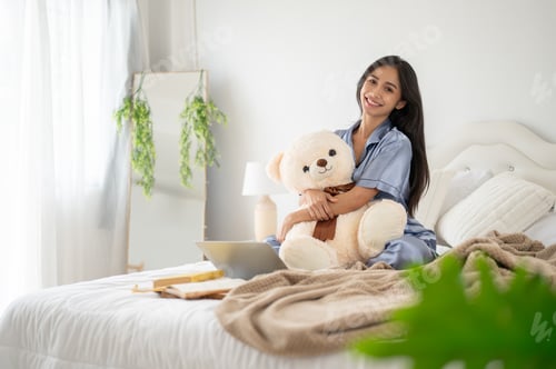 Preview: An attractive Asian woman in pajamas is hugging a teddy bear while sitting on her bed in her bedroom