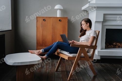 Preview: Disappointed Hispanic young woman in casual clothes sits on chair with laptop home against fireplace