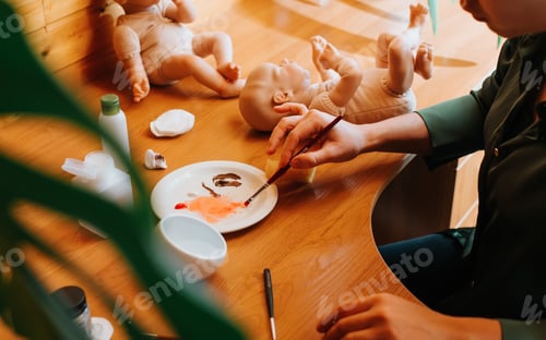 Preview: Young female craftsman decorating realistic caucasian handmade newborn baby doll sitting at table in